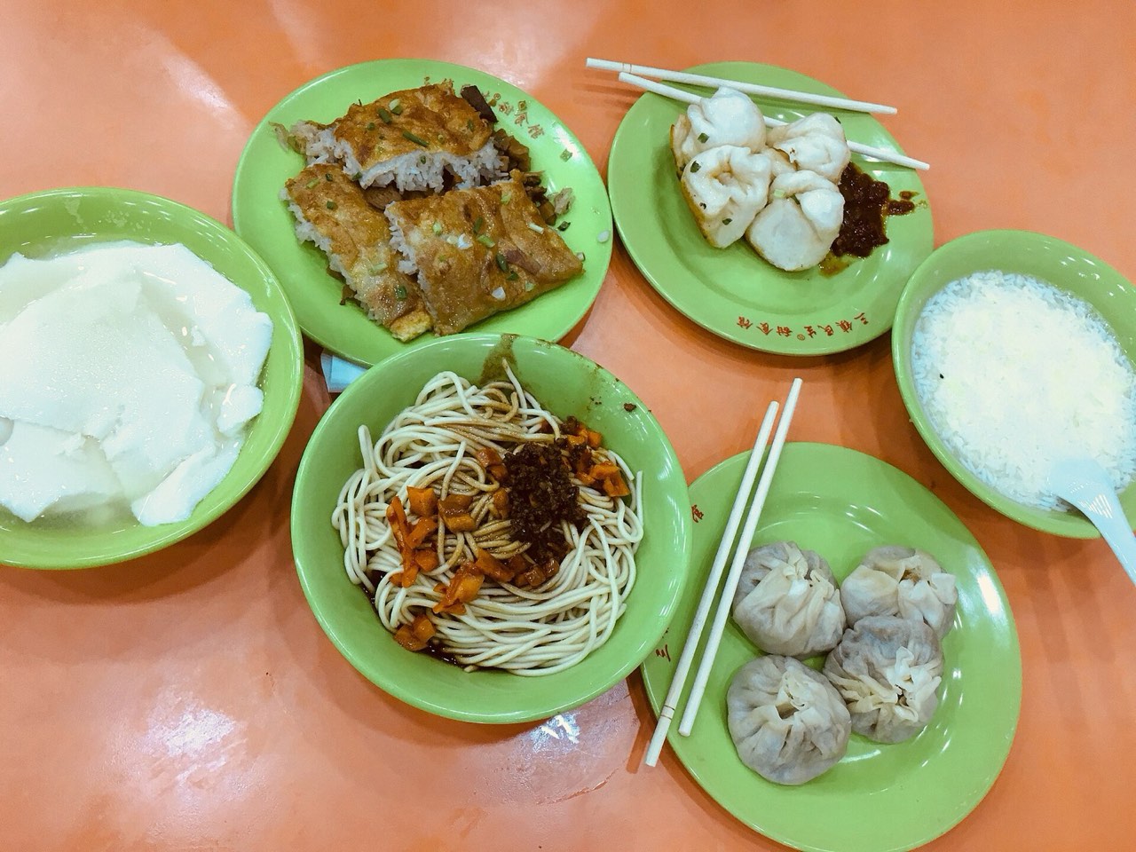 A full Wuhan guozao breakfast table: doupi, hot dry noodles, tofu pudding, shaomai, and congee in green bowls